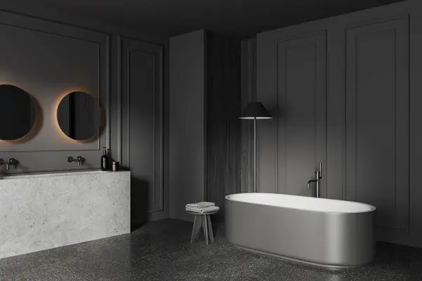 Dark home bathroom interior with bathtub and double sink, corner view side table with towel and lamp on grey concrete floor. Bathing space with minimalist furniture and molding wall. 3D rendering