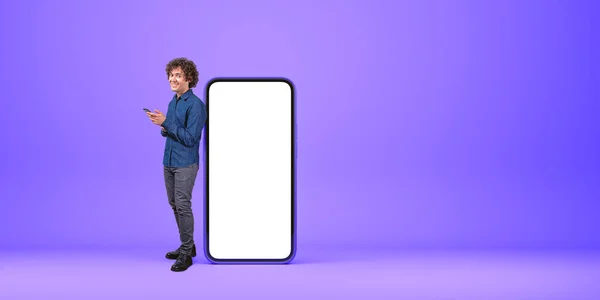 紫色の背景の上にスクリーンをモックアップして大きなスマートフォンにカーリーヘアを傾けている幸せな若者の全長の肖像画 アプリ広告とオンラインコミュニケーションの概念 コピースペース — ストック写真