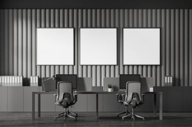 Koltukları ve bilgisayarları olan karanlık bir iş odası. İçinde belgeler olan bir büfe. Minimalist mobilyalarla birlikte çalışma alanı ve sıra sıra dizilmiş kare tuval posterleri. 3B görüntüleme