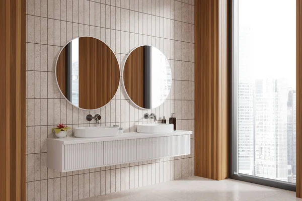 Stylish home bathroom interior with double sink and vanity, side view decor and accessories. Two washbasins and round mirror on beige tile wall, panoramic window on skyscrapers. 3D rendering