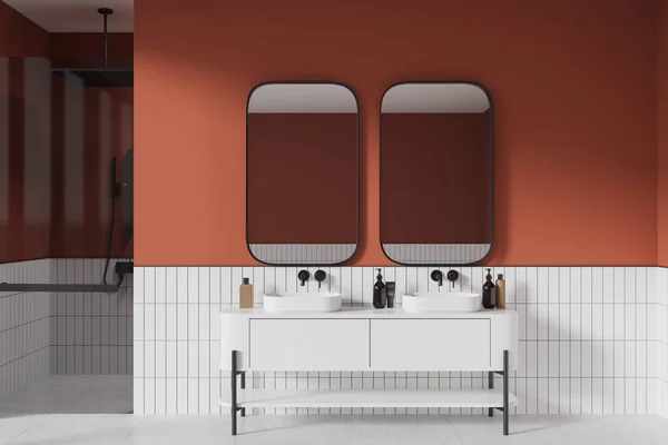 Interior of modern bathroom with orange and white tiled walls, white tiled floor, comfortable white double sink with two vertical mirrors and walk in shower with glass door. 3d rendering