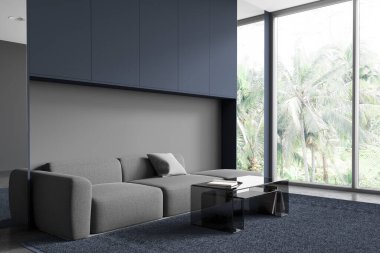 Mavi ve gri duvarlı şık oturma odasının köşesi, taş zemin, cam sehpanın yanında duran rahat gri kanepe ve tropikal manzaralı panoramik pencere. 3d oluşturma