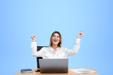 Elleri havada neşeli bir ofis kadını. Masasında dizüstü bilgisayarı ve iş belgeleriyle oturuyor. Boş mavi arkaplanı kopyalıyor. Tatil, terfi veya kâr kavramı