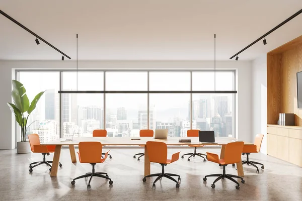 Elegant office interior with orange chairs and laptop on desk, light concrete floor. Modern meeting and coworking space, shelf with decoration and panoramic window on Kuala Lumpur. 3D rendering