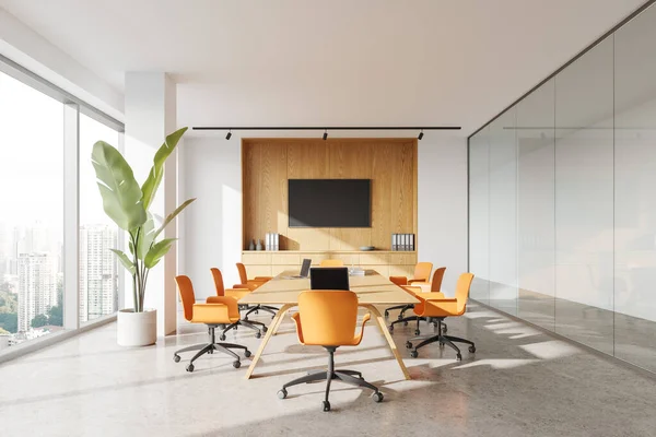 Interior of modern meeting room with white and glass walls, concrete floor, long conference table with orange chairs and flat screen TV set on the wall. 3d rendering