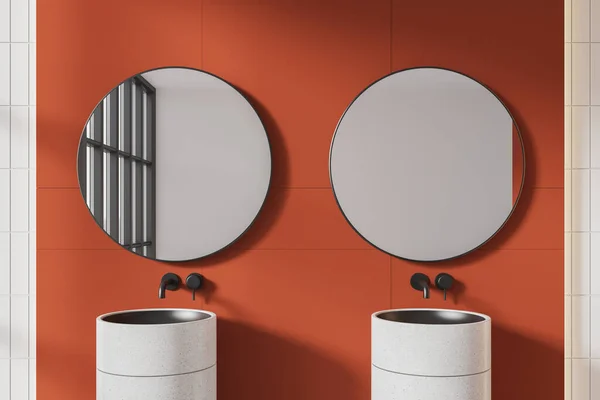 Interior of modern bathroom with bright orange and white tiled walls and comfortable white double sink with two round mirrors hanging above it. 3d rendering