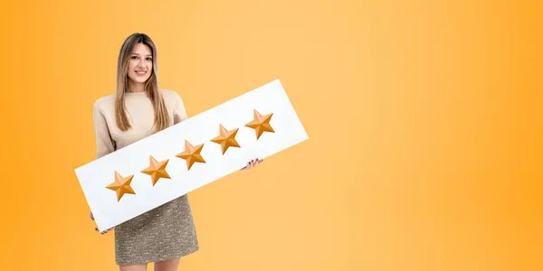 Portrait of smiling young European woman holding five star sign standing over yellow copy space background. Concept of product and service customer rating and client feedback