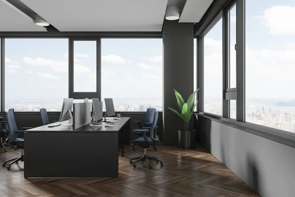Dark office interior with chairs and pc monitors on shared desk, plant on hardwood floor. Workspace with furniture and panoramic window on New York city view. 3D rendering