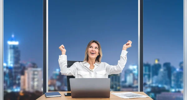 快乐的女商人举起双手 坐在写字台前 手里拿着笔记本电脑和商务文件 夜晚坐在纽约摩天大楼的全景窗前 职业发展和晋升的概念 — 图库照片