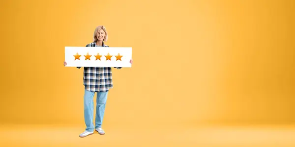 Portrait of cheerful young woman in casual clothes holding five star sign standing over orange copy space background. Concept of product and service evaluation and client feedback