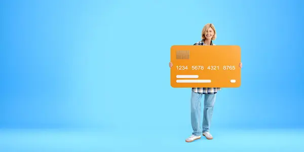 Smiling blonde woman full length showing mock up orange bank credit card, copy space empty blue background. Concept of payment, purchase, online or electronic money
