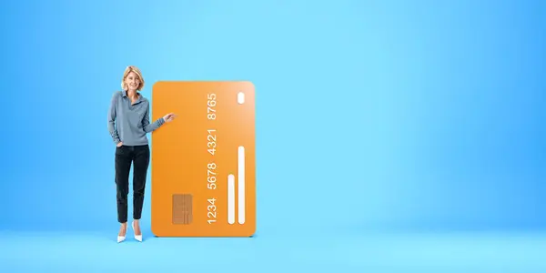 Smiling blonde woman full length finger pointing at large mock up credit card. Concept of recommendation, payment, purchase, finance and electronic money