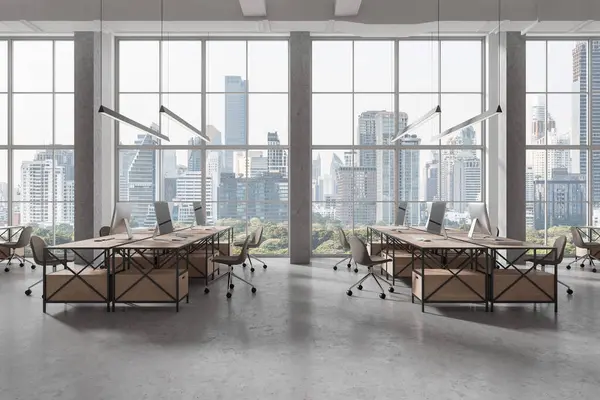 Stylish office interior with pc computers and shared desk in row, grey concrete floor. Stylish coworking space with panoramic window on Bangkok skyscrapers. 3D rendering