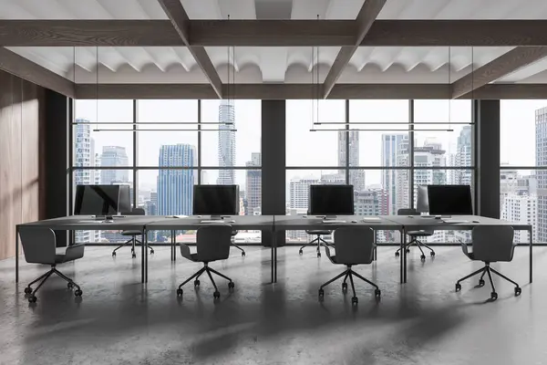 Dark office interior with seats and pc monitors, table in row on grey concrete floor. Minimalist coworking space and panoramic window on Bangkok skyscrapers. 3D rendering
