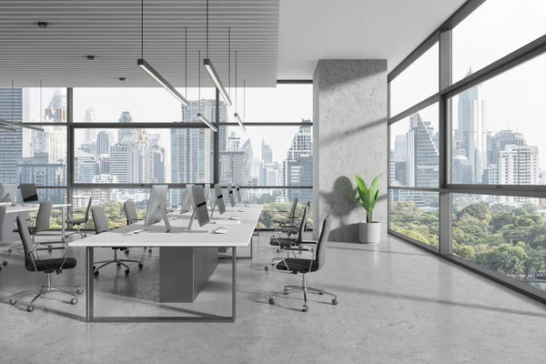 Industrial office interior with pc computers and shared desk in row, grey concrete floor. Stylish coworking space with panoramic window on Bangkok skyscrapers. 3D rendering