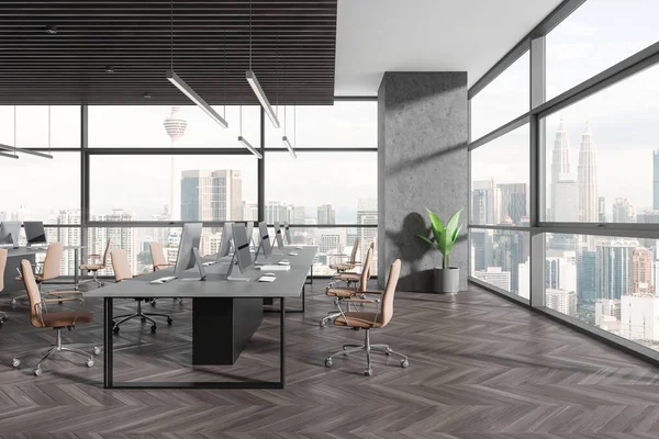 Dark coworking interior with pc desktop on table in row, hardwood floor. Stylish workplace with panoramic window on Kuala Lumpur skyscrapers. 3D rendering