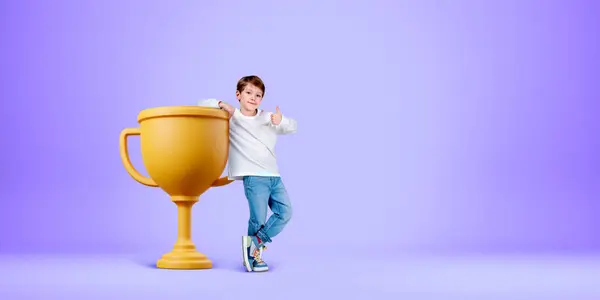 Child showing a thumb up, full length standing near big gold champion cup on light copy space purple background. Concept of first place, winner and success