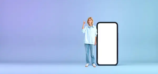 Pensive blonde woman full length standing near mockup big phone display, finger pointing up with thoughtful look. Concept of offer, recommendation, idea, start up and plan