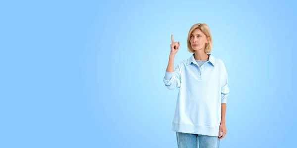 Beautiful blonde woman in long polo and denim jeans, pensive person finger point up on copy space blue background. Concept of business startup and idea