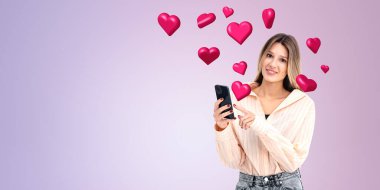 Gülümseyen bir kadın, mor bir arka planda yüzen kırmızı kalplerle çevrili akıllı telefonuna bakıyor. Aşk kavramını ya da online randevu kavramını tasvir ediyor.