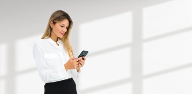Akıllı telefon kullanan güzel bir iş kadını. Boş fotokopi duvarının arka planında resmi bir portre. İş iletişimi, sosyal medya ve çevrimiçi alışveriş kavramı
