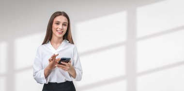Gülümseyen ve akıllı telefon kullanan, beyaz bluz giyen, pencere gölgeli beyaz bir arka plan karşısında duran bir kadın. İletişim kavramı