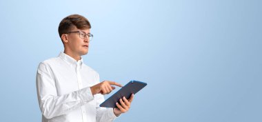 Beyaz gömlekli genç bir adam ekranı gösteren bir tablet kullanıyor. Arka plan açık mavi. Çevrimiçi eğitim ve dijital öğrenme kavramı