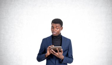 Mavi takım elbiseli ve siyah balıkçı yakalı genç bir adam tablet kullanıyor, açık gri arka planda duruyor, iş ve teknolojiyi temsil ediyor.