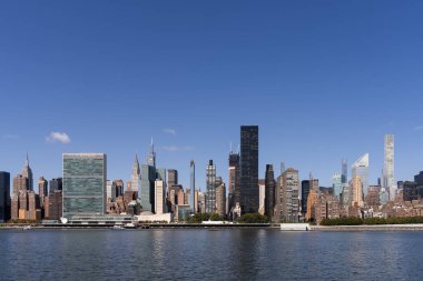 New York şehrinin ufuk çizgisi, açık mavi bir gökyüzünün altında rıhtımdaki Birleşmiş Milletler binası da dahil olmak üzere gökdelenlerin bir karışımını içeriyor. Kentsel manzara kavramı