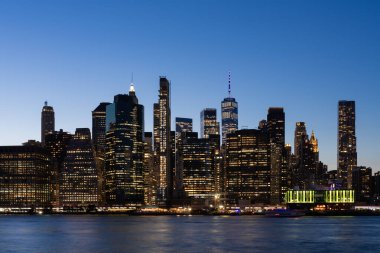 Manhattan 'ın alacakaranlıktaki ufuk çizgisinin çarpıcı görüntüsü, aydınlanmış binaların sergilenmesi, sakin bir nehir arka planında, kentsel güzellik kavramı