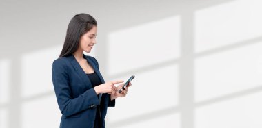 Takım elbiseli bir iş kadını akıllı telefon kullanıyor, bulanık bir arka plana karşı duruyor. Profesyonel iletişim ve teknoloji kavramı