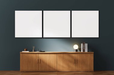 Modern, minimalist bir oda ortamında ahşap bir dolabın üzerindeki koyu renkli bir duvara asılı üç boş tuval. 3B Hazırlama