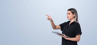 Bir elinde tablet tutan bir kadın diğerini işaret ederken, arka planda nötr bir ışığa karşı duruyor. Teknoloji ve sunum kavramı