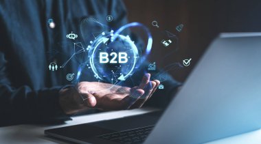 Profesyonel iş adamının elleri dijital arayüzde teknoloji simgeleriyle çevrili parlayan bir B2B simgesini tutuyor. Çevrimiçi işbirliği kavramı