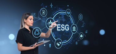 Elinde tablet tutan bir kadın, gelecekteki mavi arkaplan ile dijital arayüzde parlayan ESG simgelerine işaret ediyor. Sürdürülebilirlik ve yönetim kavramı