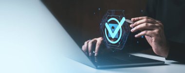 El, dizüstü bilgisayarın üzerinde parlayan bir kontrol işareti hologramıyla etkileşime geçiyor. Gradyan arka planda doğruluğu sembolize ediyor. Dijital güvenlik kavramı