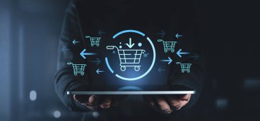 Elinde parlayan holografik alışveriş arabası simgeleri ve okları olan bir tablet tutan kişi, karanlık bir arka planda e-ticareti ve dijital perakendeciyi sembolize ediyor.