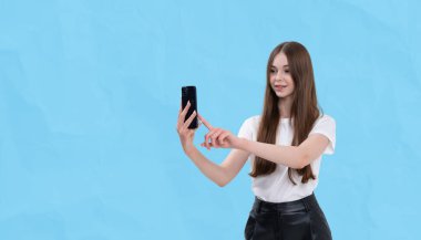 Akıllı telefon, günlük kıyafet, açık mavi arka planda izole edilmiş sosyal medya kullanımı konsepti kullanarak selfie çekerken gülümseyen genç kız