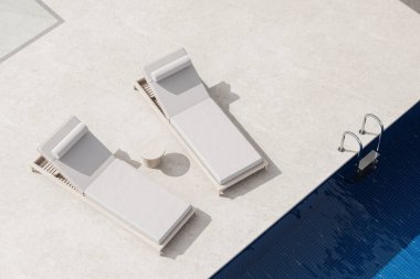 İki bej güneş şezlonglu modern havuz manzarası ve hafif taş bir verandada küçük yuvarlak bir masa, koyu mavi karo bir havuzun yanında. 3B Hazırlama