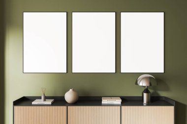 Ahşap dolabın üstündeki yeşil duvarda üç boş poster çerçevesi, içinde kitaplar, lamba ve vazo, tasarım sunumu konsepti için temiz bir düzen. 3B Hazırlama