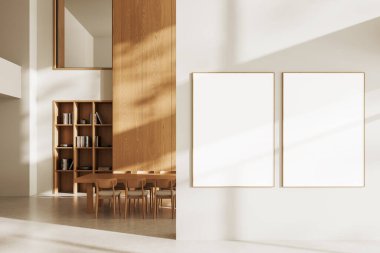 Ahşap mobilyalar, kitaplık ve güneş ışığı gölgeleriyle dolu modern bir iç mekanda beyaz bir duvara ahşap çerçeveli iki boş poster. 3B Hazırlama