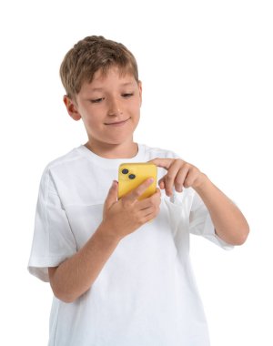 Happy child boy in white shirt holding yellow phone, touching screen with finger, standing isolated on white background, concept of digital childhood
