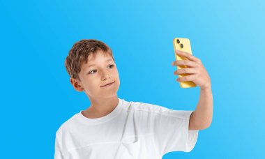 Young boy holds yellow smartphone taking selfie, realistic style, on a vibrant blue background. Concept of child and modern digital communication