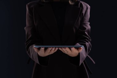Woman in formal outfit holding tablet with both hands, low light style, dark background. Concept of digital technology and modern communication