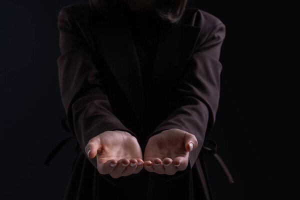 Close-up of a woman in a dark coat extending her empty hands forward in a gesture of offering or asking, photographed on a dark studio background, concept of giving