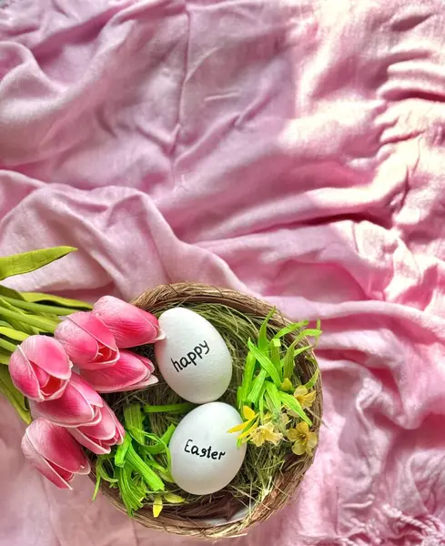 Pembe kumaşın üzerinde paskalya yumurtaları ve pembe lalelerle hasır bir yuva var.