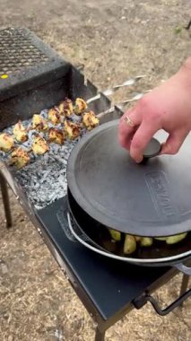 Bir el, altın patatesle dolu dökme demir bir tencerenin kapağını kaldırırken yanan kömürde kızaran et şişlerini gösteren kırsal bir açık hava yemek sahnesi..