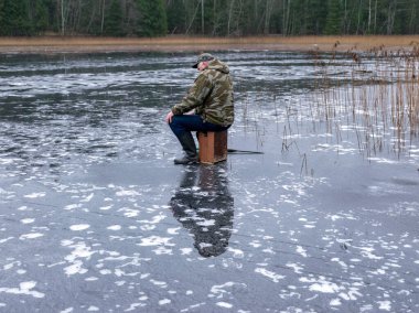 Buzda balık tutan balıkçı, buz yüzeyinde yansımalar, kıyıda çıplak ağaç siluetleri, ön planda buz dokusu.