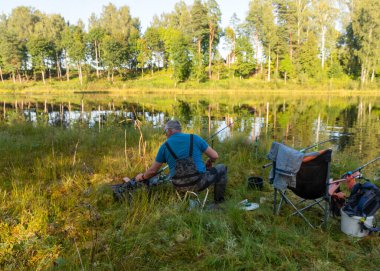 Küçük bir orman gölü olan güzel bir manzara, gölün kıyısında balıkçı, çeşitli balıkçılık özellikleri, sakin bir yaz akşamı