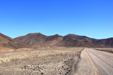 Arka planda yüksek volkanik dağlar olan çakıllı, tozlu bir yol. Jandia, Morro Jable, Fuerteventura, İspanya 'daki Kanarya Adaları